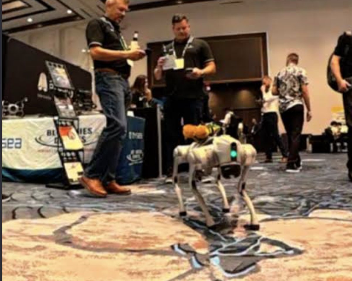 A quadruped robot moving across the floor of a hotel ballroom reception while guests look on.