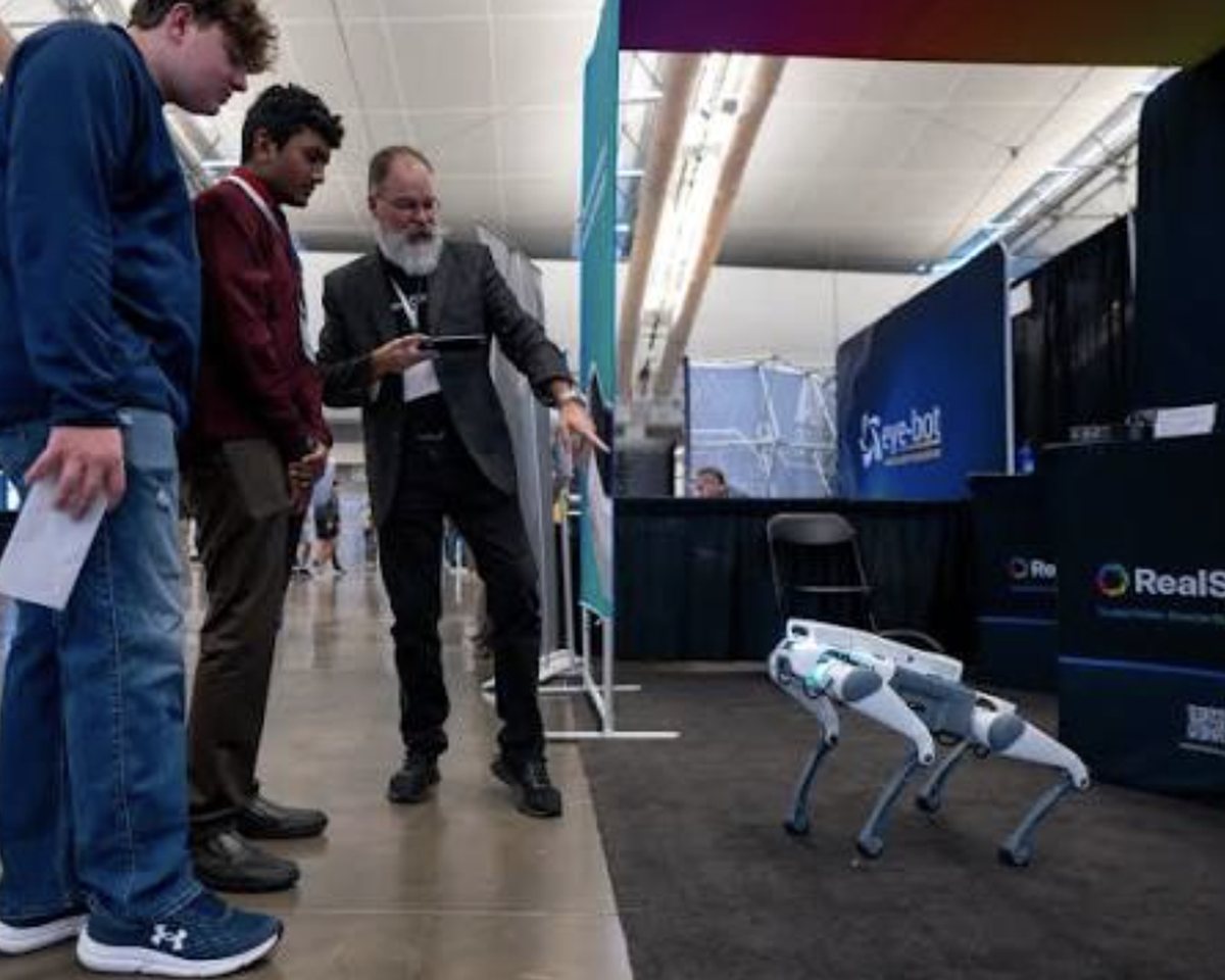 Attendees at a trade show stopping to watch a quadruped robot walk along the aisle edge.